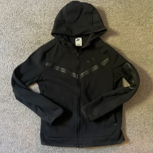 Svart Nike tech - Svart hoodiejacka från Nike med hel dragkedja och huva. Jackan har diskreta svarta detaljer över bröstet och är tillverkad i mjuk bomull och polyester. Perfekt för chill dagar eller när du vill ha en sportig look. Klassisk Nike-logga på bröstet.