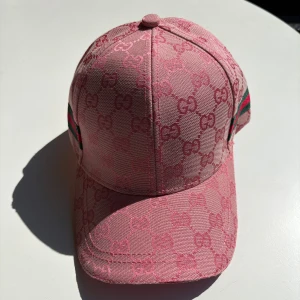 Rosa Gucci keps med GG-mönster - Snygg rosa keps från Gucci med ikoniskt GG-mönster över hela kepsen och klassiska gröna och röda ränder på sidorna. Justerbar rem baktill och insida med Gucci-logga. Tillverkad i bomull och polyester för en lyxig känsla.