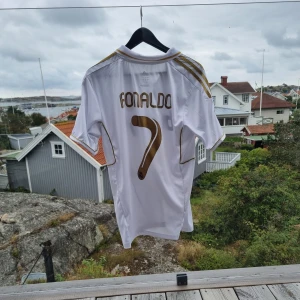 Real Madrid Ronaldo tröja Adidas - Säljer en vit Real Madrid fotbollströja från Adidas med guldiga detaljer och svarta tryck. Tröjan har korta ärmar, krage och CL-märke på ärmen. Nummer 7 och Ronaldo på ryggen, samt Bwin-logga på framsidan. Materialet är lätt och andas, perfekt för match eller träning.