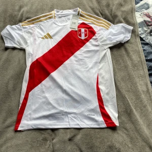 Peru landslagströja Adidas vit/röd - Snygg Peru landslagströja från Adidas i vitt med röd diagonal rand och guldiga detaljer på axlarna. Klassisk design med FPF-emblem på bröstet och AEROREADY-material som andas. Perfekt för fotbollsfans som vill sticka ut på läktaren.