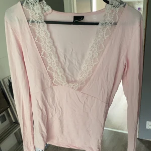 Ljusrosa långärmad topp med spets - Supersöt ljusrosa långärmad topp från Gina Tricot i storlek XS. Djup v-ringning med vit spetsdetalj längs urringningen ger en romantisk vibe. Perfekt att styla med jeans eller kjol för en feminin look.
