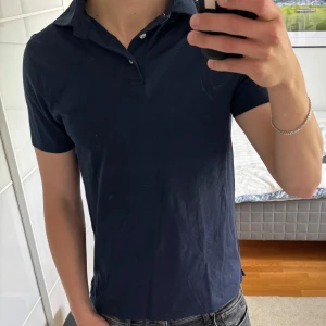 Mörkblå pikétröja Bruun & Stengade - Snygg mörkblå pikétröja från Bruun & Stengade i klassisk modell med krage och knappar framtill. Tröjan är i mjuk bomull och har korta ärmar, perfekt för en clean och stilren look. Passar dig som gillar enkel och tidlös stil. Passar även storlek S.