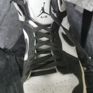 Nike Air Jordan 1 svartvita sneakers - Svartvita Nike Air Jordan 1 sneakers med klassisk hög siluett och ikoniska vingar-loggan på sidan. Skorna har svart swoosh, snörning och vit mellansula. Tillverkade i skinn med svart yttersula och perforerad tå för extra ventilation.