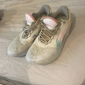 Vita sneakers med pastellfärgade detaljer - Snygga vita sneakers med pastellrosa och blå detaljer på sidan och tungan. Skorna har snörning, perforerat mönster på ovandelen och en sportig vibe. Perfekta för dig som gillar en unik och fräsch look.