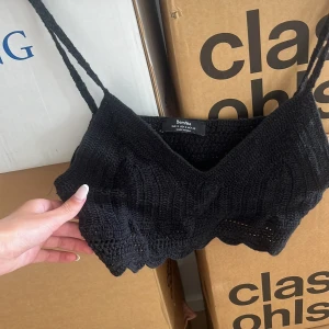 Svart virkad croppad topp Bershka - Svart virkad croppad topp från Bershka med smala axelband och vågig kant nertill. Toppen är ärmlös och har en luftig stickad känsla, perfekt för varma dagar. Enkel och stilren design som passar till många olika looks. Så fin att ha på en bikini topp nu i sommar.