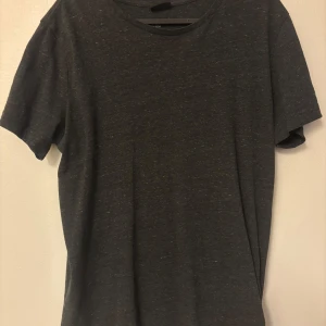 Mörkgrå basic t-shirt från H&M - En enkel och stilren mörkgrå t-shirt från H&M Basic. T-shirten har rund halsringning och korta ärmar, perfekt för en clean och avslappnad look. Tillverkad i mjuk bomull för skön komfort hela dagen.
