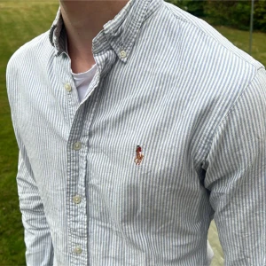 Randig skjorta från Ralph Lauren - Snygg slim fit skjorta från Ralph Lauren i ljusblått och vitt randigt mönster. Skjortan är tillverkad i 100% bomull, har klassisk krage, knäppning framtill och broderad logga på bröstet. Perfekt för en clean och stilren look. 2 små fläckar som knappt syns vet dock ej om de går bort i tvätten men för priset är de 100% värt !! Storlek 8 = XS