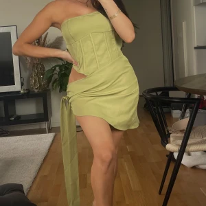 Olivgrön kort klänning PrettyLittleThing - Snygg olivgrön kort klänning från PrettyLittleThing med korsettliknande överdel, cut-out i sidan och knytband. Klänningen är axelbandslös och har en tight passform som framhäver figuren. Perfekt för dig som vill sticka ut med en trendig och unik look. Jag på bilden har normalt storlek UK4-6 därav är den för stor på mig