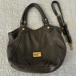 Gråbrun handväska från Marc by Marc Jacobs - Säljer en snygg grå handväska från Marc by Marc Jacobs i skinn med guldfärgade metalldetaljer. Väskan är nästan i nyskick med endast några få skrapor på vissa av metalldetaljerna. En axelrem medföljer💛