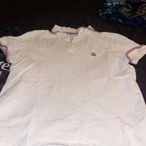 Vit Moncler pikétröja med rödblå detaljer, den är äkta. Köpte den för 1499 men säljer den för 849 nu. - Vit pikétröja från Moncler med korta ärmar och klassisk krage. Tröjan har röda och blå ränder på krage och ärmslut samt Moncler-logga på bröstet. Perfekt för en clean och sportig look. 