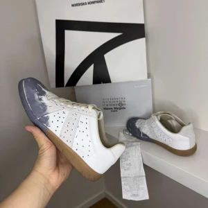 Gats, Maison Margiela - Maison Margiela sneakers i vita med blått på tån, kvitto låda och skor får man med vid köp. Skorna är 9/10 sick och är enbart lite smutsiga och repade under sulan vilket är naturligtvis meningen pris går att förhandla och skorna säljs i befintligt sick. Se sista bilden för att se kvittot. Innesulan är bytt från orginal till extra sulorna man får med när man köper dem vilket sänker priset en del!
