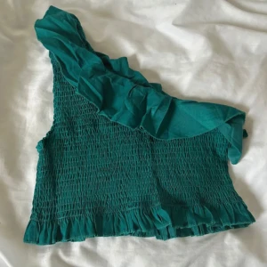 Grön one shoulder volangtopp - Snygg grön topp med one shoulder-design och volangdetalj längs axeln. Smockad över hela kroppen för en tight passform och volangkant nertill. Perfekt för dig som vill sticka ut med en cool och trendig look