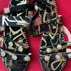 Svarta sandaler med guldmönster och pärlor - Unika sandaler i svart med abstrakt guldmönster och glittrande pärldetaljer på remmarna. Platt sula och öppen tå, perfekt för att sticka ut i sommar. Justerbara remmar och cool design som drar blickarna till sig.
