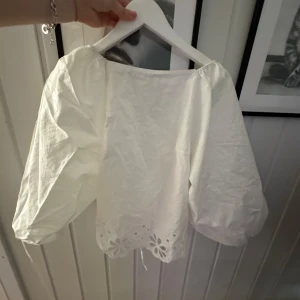 Vit blus med snörning och broderi - Superfin vit blus med puffiga ärmar och snygg snörning i ryggen. Nedtill har den broderade blommor och en scallopad kant som ger en söt vibe. Perfekt till jeans eller kjol för en trendig look.