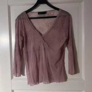 Ljuslila blus med spets från KappAhl - Superfin ljuslila blus från KappAhl med blommigt mönster i mesh och v-ringning. Blusen har spetsdetaljer längs ringningen och trekvartsärm. Tunn och luftig, perfekt för lager på lager eller som statement-piece.