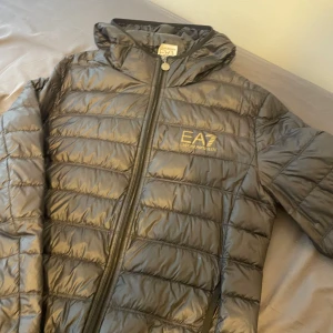 Armani jacka  - Snygg svart dunjacka från EA7 Emporio Armani med guldig logga på bröstet. Jackan har huva, dragkedja framtill och quiltad design. Perfekt för kyliga dagar och ger en clean och modern look.