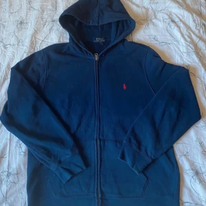 Marinblå hoodie från Polo Ralph Lauren - Snygg marinblå hoodie från Polo Ralph Lauren med dragkedja och klassisk röd logga på bröstet. Tröjan har huva, långa ärmar och är tillverkad i mjuk bomullsblandning. Perfekt för chill dagar eller när du vill ha en clean och stilren look.
