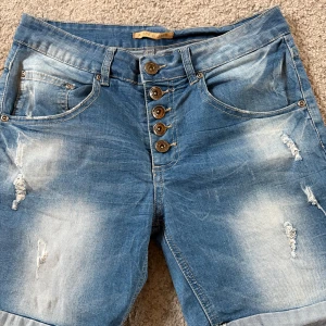 Blå jeans med slitningar och stjärndetalj - Snygga blå jeans från Place du Jour med slitna detaljer och coola slitningar framtill. Byxorna har hög midja, knäppning med flera knappar och en stjärndetalj på bakfickan. Jeansen är i klassisk denim med ljusa tvättade partier och raka ben.