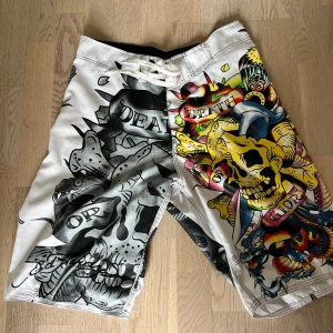 Ed Hardy shorts med dödskalletryck - Unika Ed Hardy shorts i polyester med fet dödskalle- och drakdesign, ena sidan i svartvitt och andra i färg. Snygga detaljer med snörning i midjan och coola tatueringsinspirerade motiv. Perfekta för dig som vill sticka ut på stranden eller festivalen.