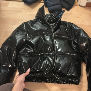 Svart glansig pufferjacka PLT - Säljer en svart, glansig pufferjacka från PrettyLittleThing. Jackan har hög krage, dragkedja framtill och en riktigt fet, vadderad look. Perfekt för dig som vill sticka ut med en edgy vibe i vinter. Jackan är i syntetmaterial och har lång ärm.