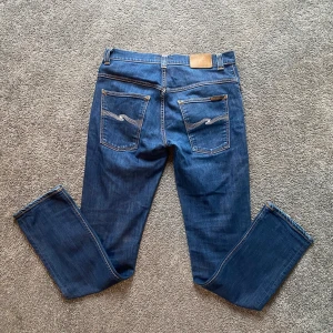 Nudie Jeans Thin Finn blå jeans 34/34 - Säljer ett par klassiska blå Nudie Jeans Thin Finn i storlek 34/34. Jeansen har smal passform, snygga bakfickor med vågig söm och är gjorda i 98% ekologisk bomull och 2% elastan för extra stretch. Perfekta för dig som gillar stilrena och bekväma jeans.