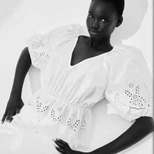 Vit blus med broderade detaljer - Superfin vit blus med v-ringning, puffiga halvlånga ärmar och broderade håldetaljer. Blusen har resår i midjan för en snygg siluett och volangkant nertill. Perfekt för dig som gillar romantisk och luftig stil. Slutsåld på hemsidan 