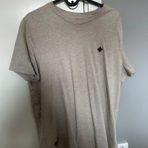 Beige Morris t-shirt  - Snygg beige t-shirt med korta ärmar och rund hals. På bröstet finns en liten svart broderad detalj som ger en clean och stilren look. Perfekt basic-plagg i mjukt material som passar till allt.