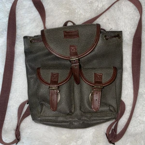 Khaki ryggsäck med bruna detaljer - Snygg grå ryggsäck med bruna skinnremmar och två framfickor med spännen. Väskan har justerbara axelband i tyg och lock med spänne. Perfekt för dig som vill ha en stilren och praktisk väska med vintagekänsla.
