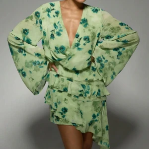  omlottklänning med volanger - Superfin ljusgrön kortklänning med blågröna blommor, djup v-ringning och långa vida ärmar. Klänningen har omlottliv och flera lager volanger i kjolen som ger en flowy känsla. Perfekt för dig som vill sticka ut med en romantisk och trendig look.