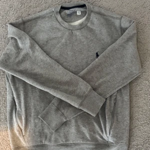 Grå sweatshirt från Polo Ralph Lauren - Klassisk grå sweatshirt från Polo Ralph Lauren med rund halsringning och broderad marinblå logga på bröstet. Tröjan har långa ärmar, ribbade muddar och är tillverkad i mjuk bomullsmix. Perfekt för en chill och stilren look.