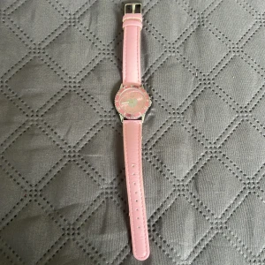 Rosa klocka med hjärta från Regal - Söt klocka från Regal med ljusrosa läderarmband och silverfärgat spänne. Urtavlan är rosa med ett glittrigt hjärta i mitten och tydliga siffror. Boetten är i silverfärgad metall och klockan har en klassisk rund form. Perfekt accessoar för dig som gillar rosa och detaljer.