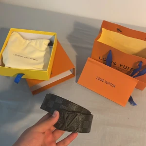 Louis Vuitton svart rutigt skärp - Säljer ett exklusivt Louis Vuitton bälte i svart skinn med klassiskt rutigt mönster och stor LV-logga i metall framtill. Bältet kommer med originalbox, dustbag och kvitto. Perfekt accessoar för att lyfta din outfit med lyxig känsla.