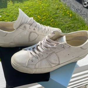 Vita sneakers från Philippe Model - Snygga vita sneakers från Philippe Model Paris med klassisk låg profil och rund tå. Skorna är i skinn med diskreta detaljer och logotyp på sidan. De har platt sula och snörning framtill, perfekt för en clean och stilren look.