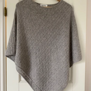 Grå kashmirponcho från Davida - Supermjuk och lyxig grå poncho från Davida i 100% kashmir. Tröjan har ett  kabelstickat mönster, rund hals och avslappnad passform. Jag säljer även en likadan i rosa.