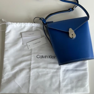 Blå axelväska från Calvin Klein - Snygg blå axelväska från Calvin Klein i stilren bucket-modell. Väskan har justerbar rem, vita kontrastsömmar och silverfärgat lås framtill. Tillverkad i slätt skinn och levereras med original dustbag och tags. Låset kan va lite svårt att öppna ibland men den fungerar🩷pris går att diskuteras