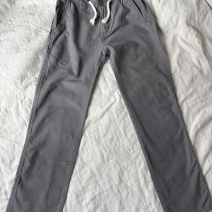 Grå linnebyxor från Lindex med snörning - Mörkgrå linnebyxor från Lindex i storlek 158. Byxorna har elastisk midja med vit snörning, raka ben och sidfickor fram.