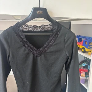 Svart långärmad topp med spets - Svart långärmad tajt topp  med snygga spetsdetaljer vid v-ringad hals och ärmslut. Mjuk och stretchig bomullskänsla, perfekt för dig som gillar en stilren look med lite extra edge. Passar till både jeans och kjol.