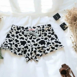 Svartvita shorts med ko-mönster och rosett - Superfina shorts med svartvitt ko-mönster och en söt ljusgrå rosett framtill. De har volangkant nedtill och elastisk midja för extra komfort. Perfekta för dig som gillar unika och lekfulla plagg.