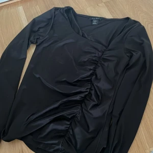Svart långärmad topp från New Yorker  - Snygg svart långärmad topp från New Yorker med cool rynkdetalj längs framsidan. Toppen har en lätt glansig finish och figurnära passform, perfekt för att lyfta din outfit. Materialet känns mjukt och stretchigt mot huden.