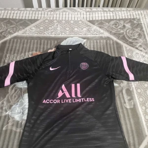 Svart PSG långärmad fotbollströja Nike - Svart långärmad fotbollströja från Nike och Paris Saint-Germain med rosa detaljer och logga på bröstet. Tröjan har halv dragkedja framtill och subtilt mönster i tyget. Materialet är lätt och andas, perfekt för träning eller match.
