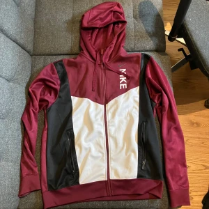 Nike vinröd hoodie med dragkedja - Snygg hoodie från Nike i vinrött, svart och vitt. Tröjan har huva med snörning, hel dragkedja framtill och två fickor med dragkedja. Tillverkad i mjukt syntetmaterial, perfekt för chill eller träning. Nike-logga på bröstet och sportig design.