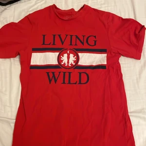 Röd t-shirt Pull&Bear Living Wild S - Röd t-shirt från Pull&Bear med trycket 'LIVING WILD' och ett lejonmotiv framtill. T-shirten har korta ärmar, rund hals och är gjord i mjuk bomull. Snygg kontrast med vita och mörkblå detaljer över bröstet. Perfekt för en chill och trendig look.
