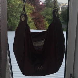 Vinröd axelväska i sammet från NYPD - Säljer en vinröd axelväska från NYPD i mjuk sammet med detaljer i skinnimitation. Väskan har guldfärgad dragkedja, innerfoder med mönster och ett snyggt metallmärke framtill. Perfekt storlek för det viktigaste och riktigt trendig look.