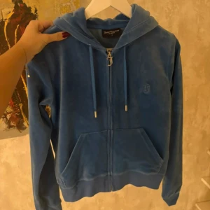 Blå hoodie Juicy Couture S - Blå hoodie från Juicy Couture i mjuk velour med dragkedja och huva. Tröjan har långa ärmar, snörning i huvan och fickor framtill. Ikonisk J-dragkedja och broderad logga på bröstet.