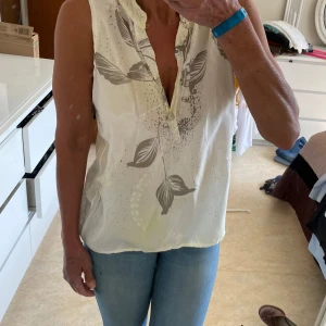 Vit -gul blus med grått bladmönster - Säljer en luftig vit +gul blus med grått bladmönster och v-ringning. Blusen är ärmlös och har en knapp framtill vid halsen. Perfekt för dig som gillar en avslappnad och fräsch stil. Materialet känns lätt och svalt, perfekt för varmare dagar.