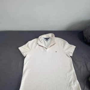 Vit pikétröja från Tommy Hilfiger - Klassisk vit pikétröja från Tommy Hilfiger med liten broderad logga på bröstet. Tröjan har korta ärmar, krage och knappar framtill. Tillverkad i mjuk bomull som känns skön mot huden. Perfekt för en clean och stilren look. Använd 4 gånger. 