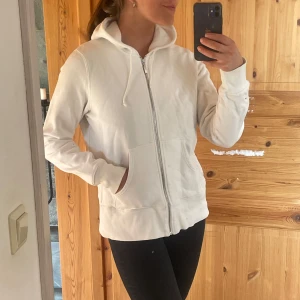 Vit hoodie med dragkedja och huva - Säljer en stilren vit hoodie med dragkedja framtill och huva med snörning. Tröjan har långa ärmar, ribbade muddar och två fickor framtill. Perfekt för en avslappnad look och enkel att matcha med det mesta.