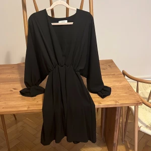 Svart omlottklänning från Chiquelle - Svart kortklänning från Chiquelle med djup v-ringning och omlottkänsla. Klänningen har långa, lösa ärmar och markerad midja med snygg rynkdetalj. Perfekt för dig som gillar stilrena och trendiga plagg med en relaxed vibe.