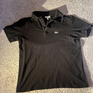 Svart pikétröja från Lacoste - Klassisk svart pikétröja från Lacoste. Perfekt till sommaren storlek 164 för 14 åringar.