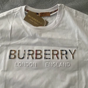 Vit Burbery t shirt  - Vit t-shirt från Burbery med klassisk rund hals och korta ärmar. Stor 3D-logga framtill med Burberry-text i märkets ikoniska rutiga mönster och texten 'London England' under. Snygg och stilren design, perfekt för dig som gillar exklusiva märken.